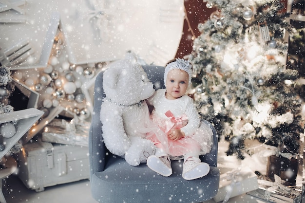 Retrato de lindo bebé en perspectiva hermosa sentado en un sillón con osito de peluche blanco y sonriendo a la cámara. Interior decorado de Navidad y árbol iluminado.