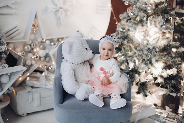 Retrato de lindo bebé en perspectiva hermosa sentado en un sillón con osito de peluche blanco y sonriendo a la cámara. Interior decorado de Navidad y árbol iluminado.