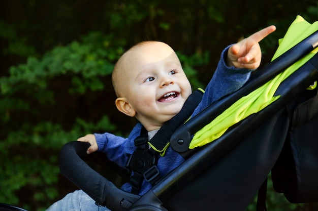 Un retrato de un lindo bebé sonriente feliz sentado en el cochecito mirando hacia un lado y apuntando con el dedo hacia arriba en el parque durante la caminata en verano.