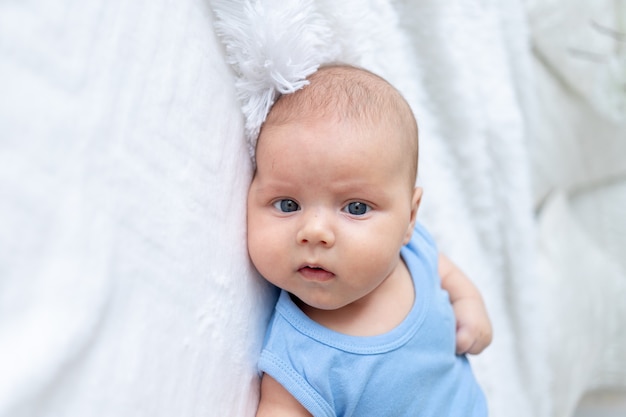 Retrato de un lindo bebé de tres meses en un traje azul sobre una cama blanca en casa