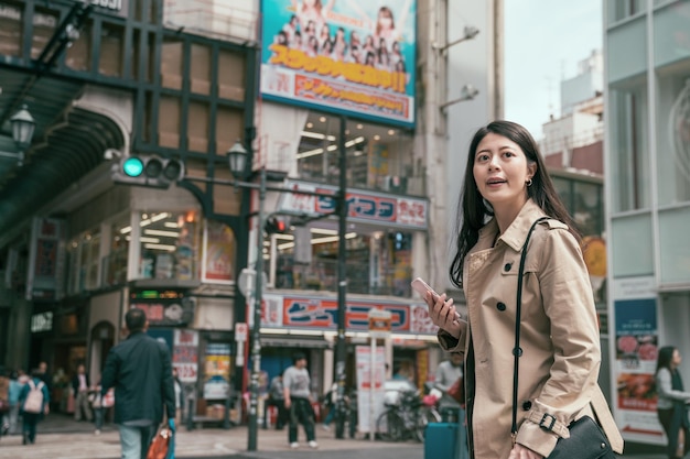 retrato lindo jovem asiático japonês gerente segurando o telefone inteligente na mão e olhando para trás, encontrando a pessoa. senhora paciente esperando namorado no encontro depois do trabalho. menina fazendo compras em shinsaibashi