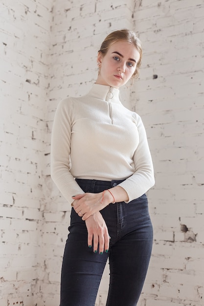 Retrato de lindo modelo adolescente pensativo con suéter blanco y jeans negros. Mujer joven flaca caucásica con cabello rubio se encuentra frente a la pared de ladrillo blanco. Señora bonita natural de pie en el estudio