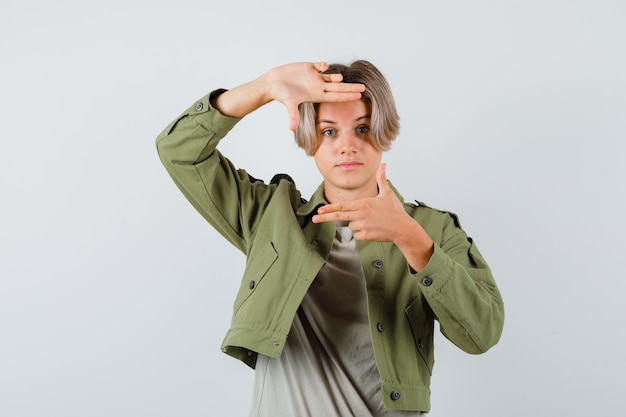 Retrato de lindo muchacho adolescente mostrando gesto de marco con pistolas de dedo en chaqueta verde y mirando confiado vista frontal