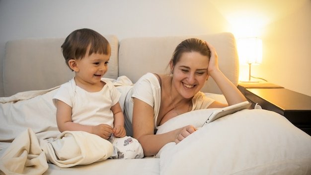 Retrato de lindo niño acostado con la madre en la cama y viendo videos en tableta digital.