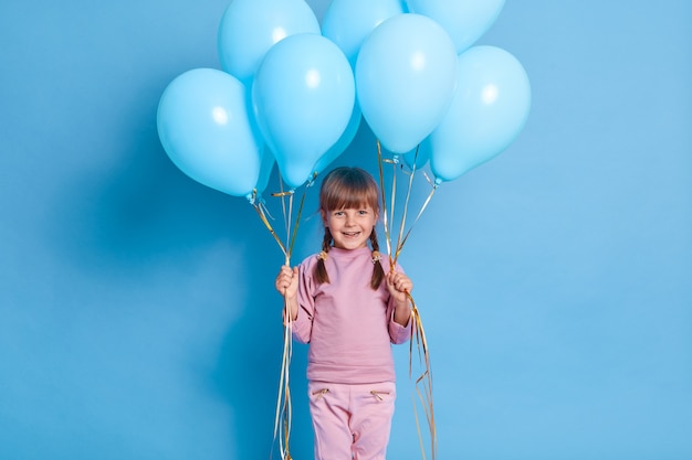 Retrato de lindo niño en edad preescolar posando contra la pared azul con globos