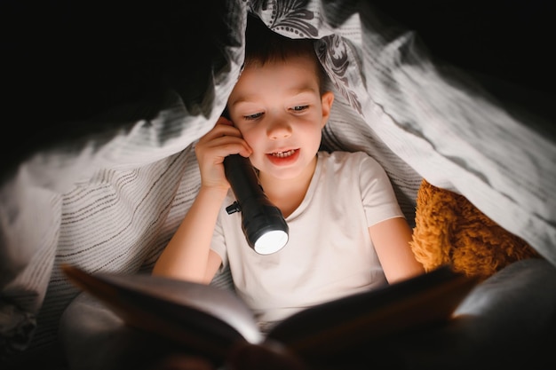 Retrato de un lindo niño leyendo en la cama con una linterna en una habitación oscura, disfrutando de los cuentos de hadas