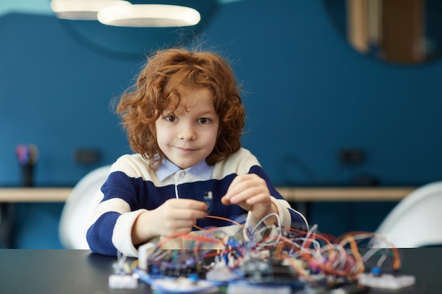 Retrato de lindo niño de pelo rizado robot de construcción en clase de ingeniería y mirando a cámara, espacio de copia