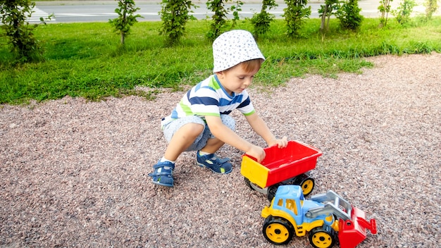 Retrato de lindo niño pequeño de 3 años sentado en el patio de recreo en el parque y jugando con coloridos camiones de juguete de plástico