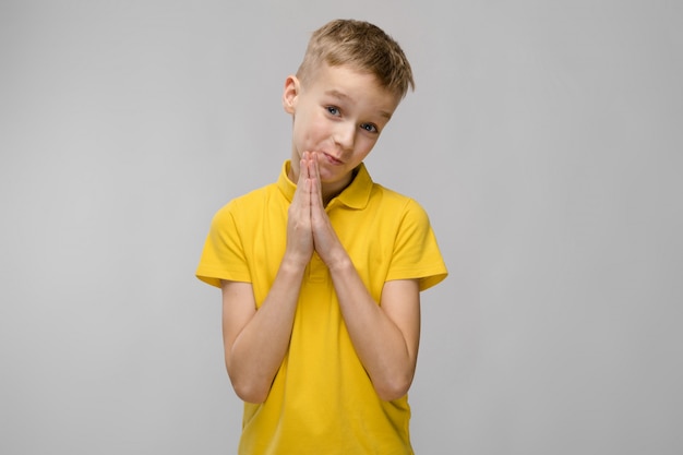 Retrato de lindo niño pequeño rubio caucásico en camiseta amarilla con la esperanza de pedir perdón en gris