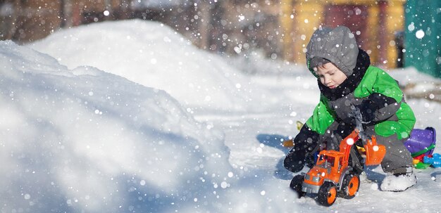 Retrato de lindo niño pequeño sentado en la nieve y jugando con su juguete tractor amarillo en el parque. Niño jugando al aire libre