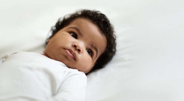 Retrato de un lindo y rizado bebé recién nacido afroamericano yace en una cama blanca y suave en el dormitorio