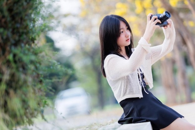 Retrato lindo sonríe asiático de una atractiva joven adolescente sosteniendo la cámara en florecientes flores amarillas doradas de Tabebuia Chrysotricha con el parque en primavera en el fondo de la noche en Tailandia