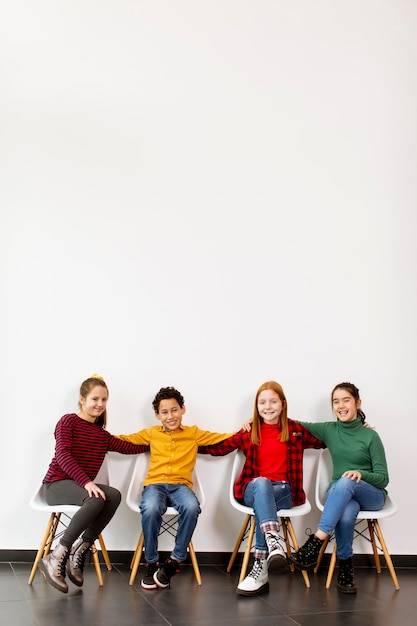 Retrato de lindos niños pequeños en jeans sentados en sillas contra la pared blanca