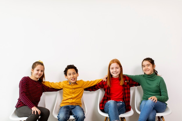 Retrato de lindos niños pequeños en jeans sentados en sillas contra la pared blanca