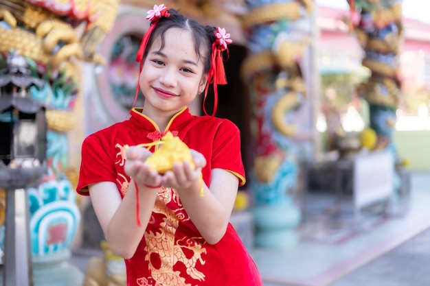 Retrato lindos sorrisos menina asiática bonitinha vestindo vermelho tradicional chinês cheongsam decoração foco mostrar saco de dinheiro dourado para o festival do ano novo chinês no santuário chinês na tailândia