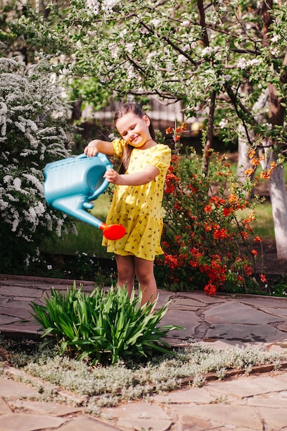 Retrato de longitud completa de estilo de vida de una niña sonriente en un vestido amarillo regando flores de un jardín ...