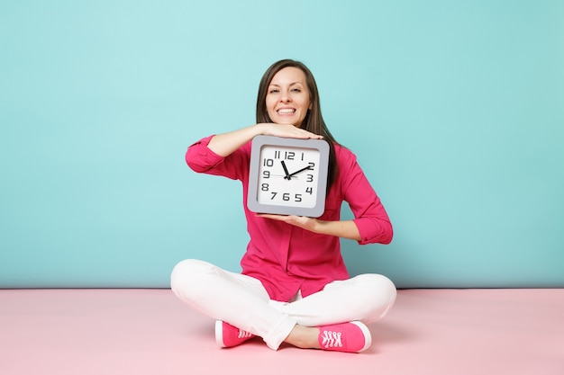 Retrato de longitud completa mujer joven en camisa rosa blusa pantalón blanco sentado en el piso mantenga el reloj