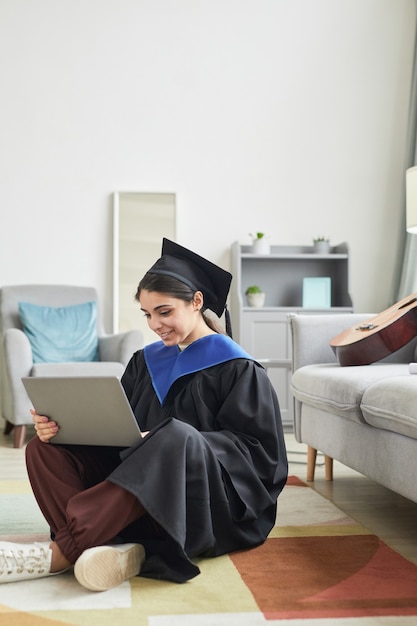 Retrato de longitud completa vertical de mujer joven con toga de graduación y usando la computadora portátil mientras está sentado en el piso en casa