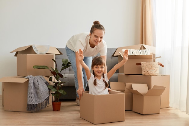 Retrato de madre e hija felices divirtiéndose durante la mudanza a un apartamento nuevo, expresando optimismo, niña sentada en una caja de cartón con los brazos levantados, reubicación, jugando con la madre.