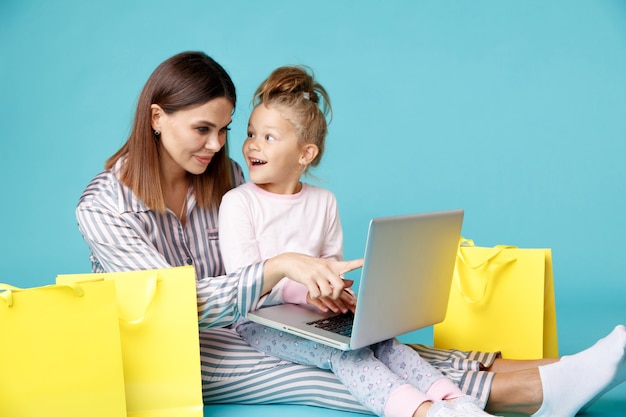 Retrato de madre e hija con laptop sentada en el piso aislado en la habitación azul. Concepto de compras familiares en línea.