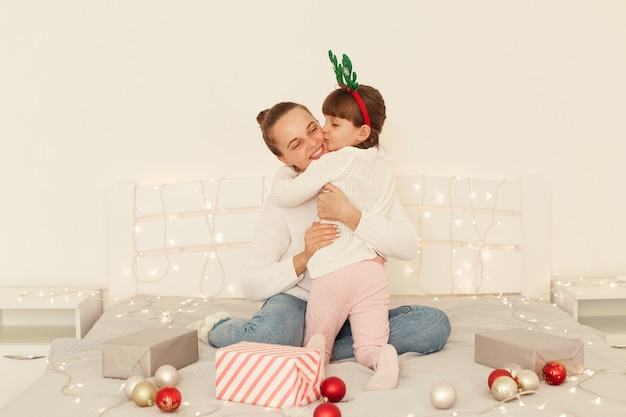 Retrato de madre e hija optimistas con suéteres blancos de estilo casual sentados en la cama, niño con cabello oscuro y coletas besando la mejilla de mamá, felicitando con año nuevo.