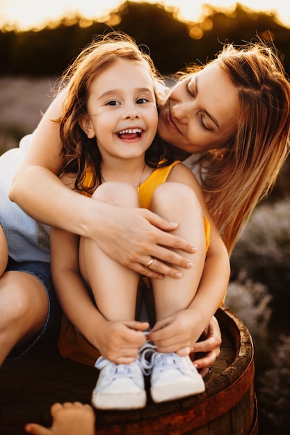 Retrato de una madre encantadora y su hija mientras la niña está mirando a la cámara riendo mientras la madre la abraza desde atrás y la mira contra el atardecer.