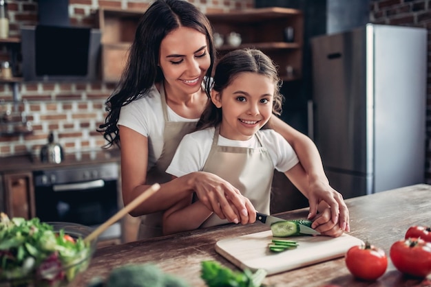 Retrato de madre feliz enseñando a su hija a cortar pepino para la ensalada en la cocina