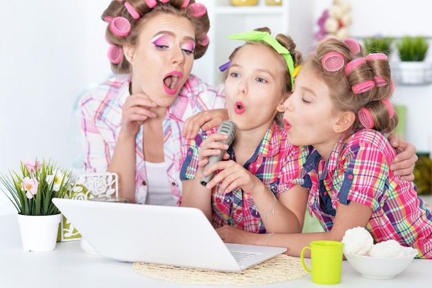 Retrato de madre feliz y pequeñas hijas en rizadores de pelo con portátil en casa