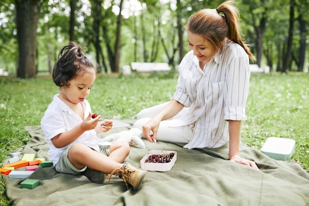 Retrato de la madre joven feliz con el niño pequeño lindo en el parque que se sienta en la hierba verde ...