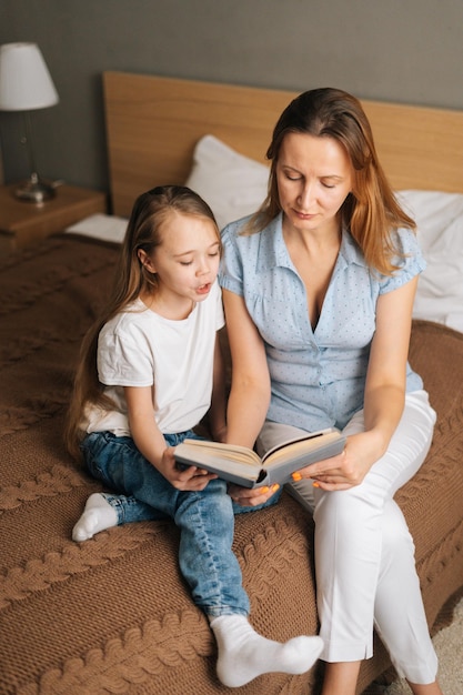 Retrato de una madre joven y una hija adorable leyendo libros para niños juntos sentados en la cama en el dormitorio de la guardería. Concepto de actividad de ocio familiar en casa.