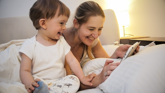 Retrato de madre riendo feliz con hijo pequeño acostado en la cama por la noche y con tablet PC.