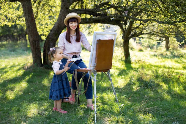 Retrato de madre sonriente joven, mostrando a su pequeña hija pintando un cuadro en el caballete, divirtiéndose juntos y pasando su tiempo libre al aire libre en el parque o jardín verde de la primavera. Copia espacio