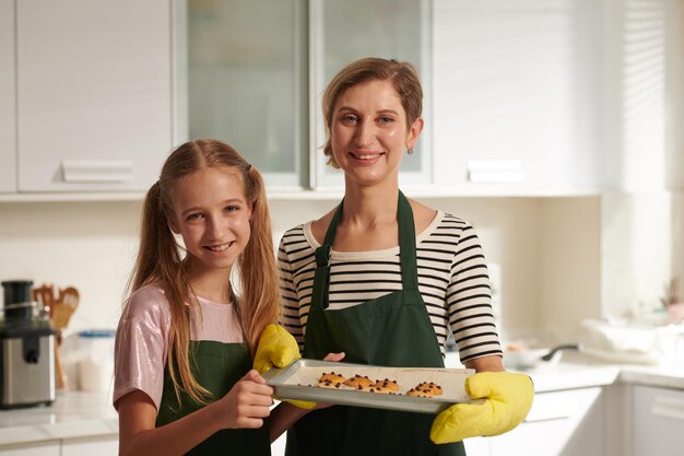 Retrato de una madre sonriente y su hija adolescente sosteniendo una bandeja con galletas que hornearon juntas