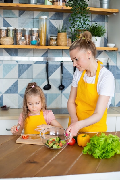Retrato de madre con su hija en delantales amarillos preparando una ensalada saludable para el almuerzo.