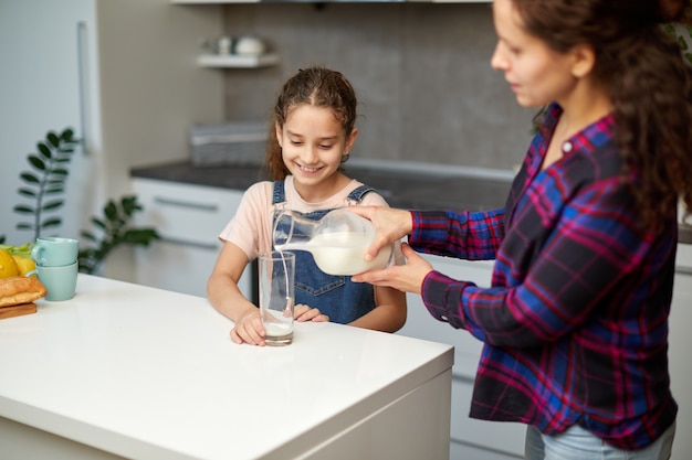 Retrato de una madre vierte su linda hija sonriente leche en vaso para el desayuno.