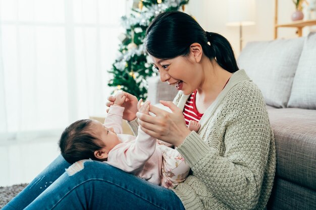 retrato mãe chinesa sentado no sofá está interagindo com seu bebê feliz. muito jovem mãe está segurando as mãos da menina e brincando com ela. momentos reais