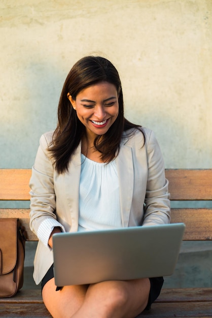 Retrato de una maestra latina sentada en un banco y trabajando felizmente con su computadora en el espacio de copia de la calle