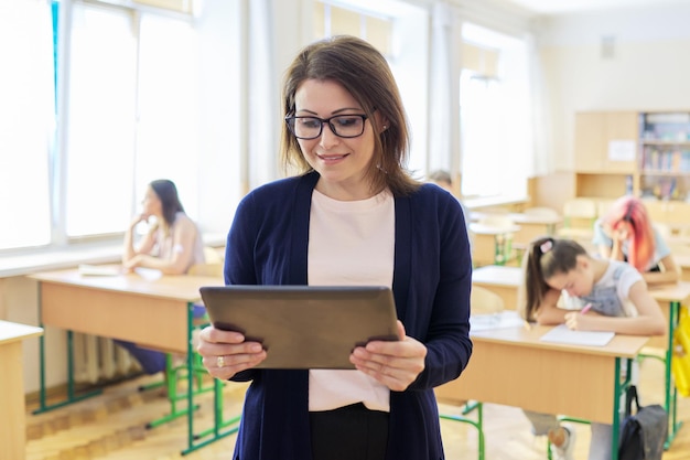 Retrato de una maestra madura en el aula con una tableta digital, una mujer sonriente y segura de sí misma en el fondo del aula con estudiantes adolescentes