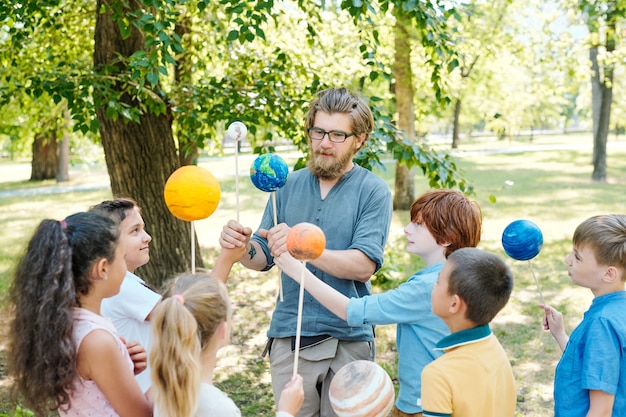 Retrato de maestro calvo apuntando al modelo de planeta y sonriendo mientras habla con un grupo de niños durante la clase al aire libre en la luz del sol