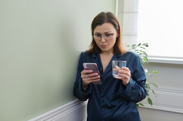 Retrato de mañana de hermosa mujer madura con vaso de agua y smartphone