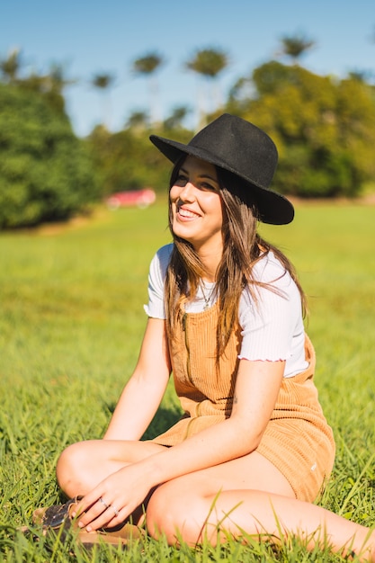 Retrato de maravilloso modelo femenino blanco con sombrero sentado en el campo