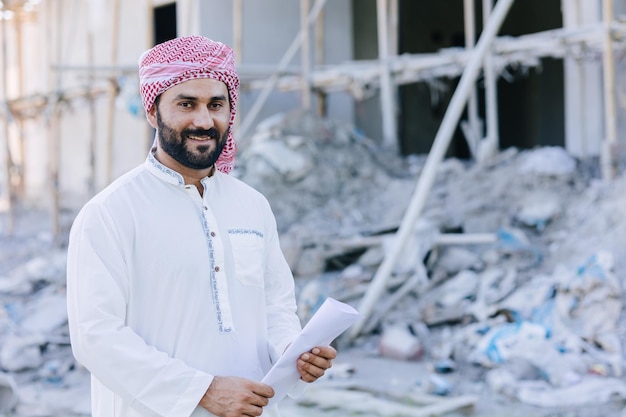 Retrato masculino de negócios de engenheiro adulto muçulmano árabe em pé com planta baixa no canteiro de obras