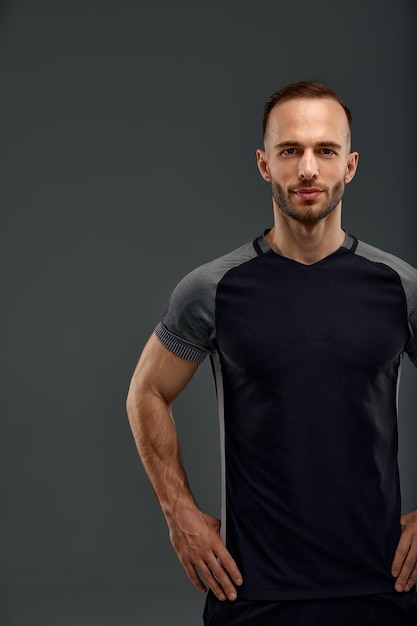 Retrato masculino de estudio de un apuesto y poderoso modelo atlético con un cuerpo musculoso saludable en una camiseta negra de moda que se burla de las poses sobre un fondo gris