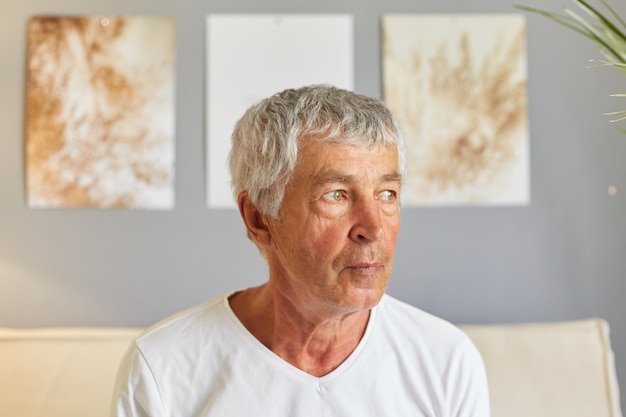 Retrato de masture hombre jubilado mirando a un lado abuelo anciano con expresión facial tranquila hombre con camiseta blanca sentado en el sofá en el interior de la casa