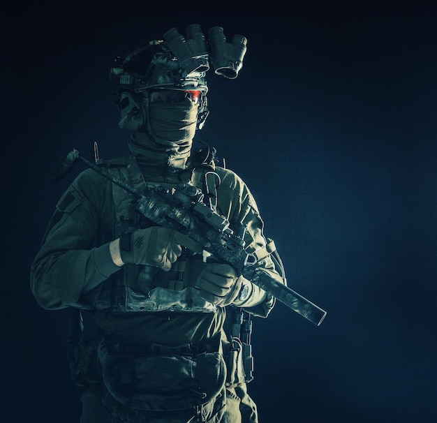 Retrato de media longitud de bajo perfil de un mercenario profesional de combate de comando de élite que oculta su identidad detrás de gafas de máscara de pie en la oscuridad con una mini ametralladora en las manos usando un dispositivo de visión nocturna