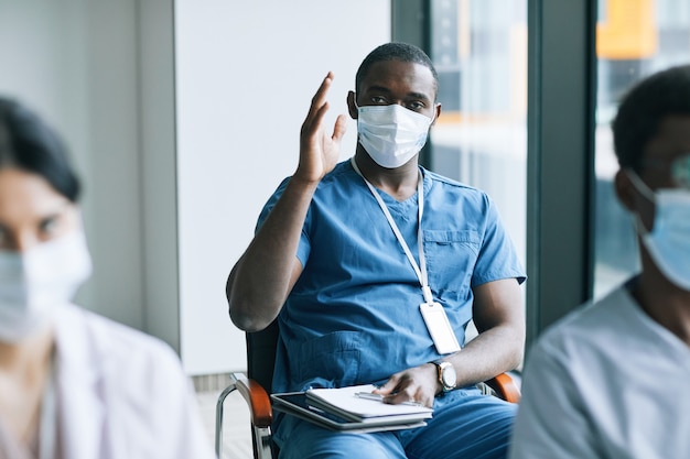 Retrato de médico afroamericano levantando la mano para hacer preguntas durante el seminario o conferencia, espacio de copia