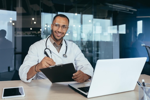 Retrato de un médico afroamericano que trabaja dentro de la oficina de una clínica moderna en la mesa usando una computadora portátil