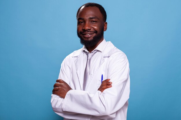 Retrato de un médico afroamericano sonriente posando con los brazos cruzados en el estudio. Residente médico profesional que parece confiado de pie en una bata blanca de laboratorio. Profesional sanitario.