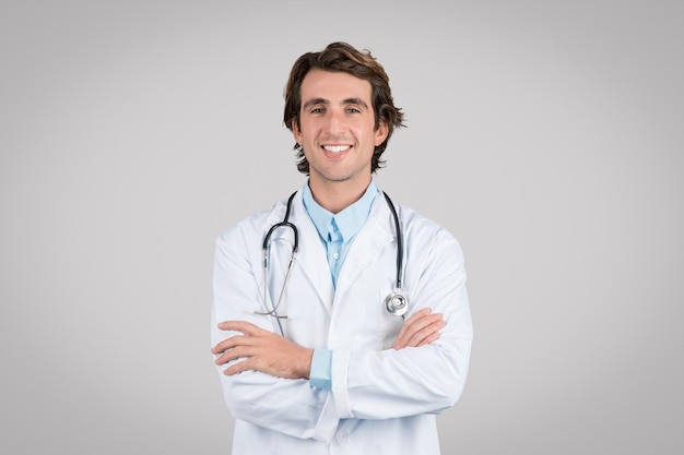 Retrato de un médico alegre posando con los brazos cruzados y sonriendo a la cámara aislado sobre el gris