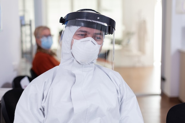 Retrato de médico cansado en el consultorio dental mirando a la cámara vistiendo un protector facial y general sentado en una silla en la sala de espera de la clínica. Concepto de nueva visita normal al dentista en caso de brote de coronavirus.